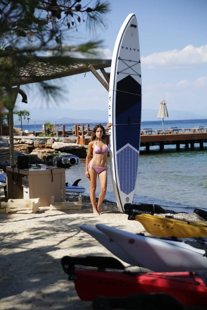Altın yazın tadını çıkaran Bodrum sahilinde, Volvo logolu paddleboard'a yaslanmış, pembe bikinili bir kadın, arkasında masmavi deniz, iskele ve diğer su sporları ekipmanlarıyla güneşli bir günün keyfini çıkarıyor.