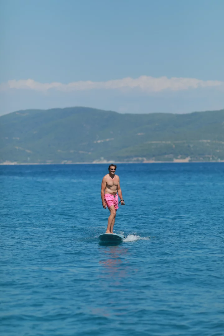 Masmavi ve berrak denizde pembe şortlu, üstsüz, güneş gözlüklü bir adam sörf tahtası üzerinde dengede durarak keyifli bir Bodrum plaj gününün tadını çıkarıyor; arka planda yemyeşil dağlar ve açık mavi gökyüzü var.