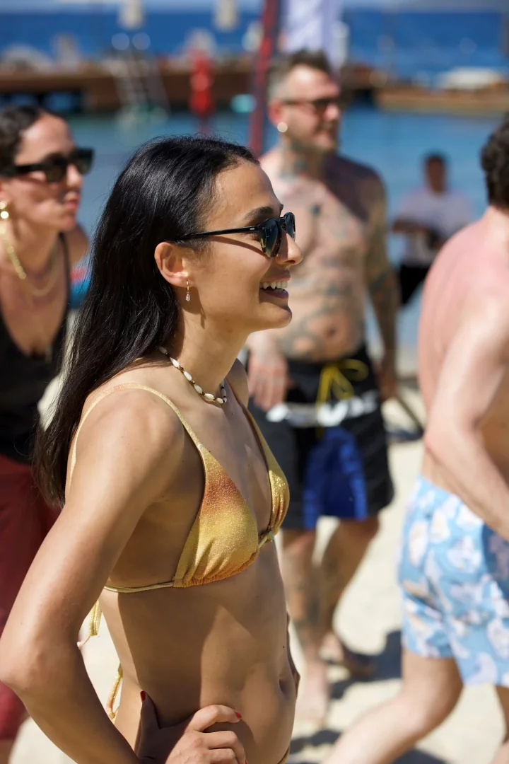 Koyu saçlı, güneş gözlüklü genç bir kadın, parıldayan altın rengi bir bikini üstü ve deniz kabuğu kolyesiyle güneşli bir Bodrum plajında neşeyle gülümsüyor, arkasında diğer plaj ziyaretçileriyle Altın Yaz'ın keyifli atmosferini yansıtıyor.