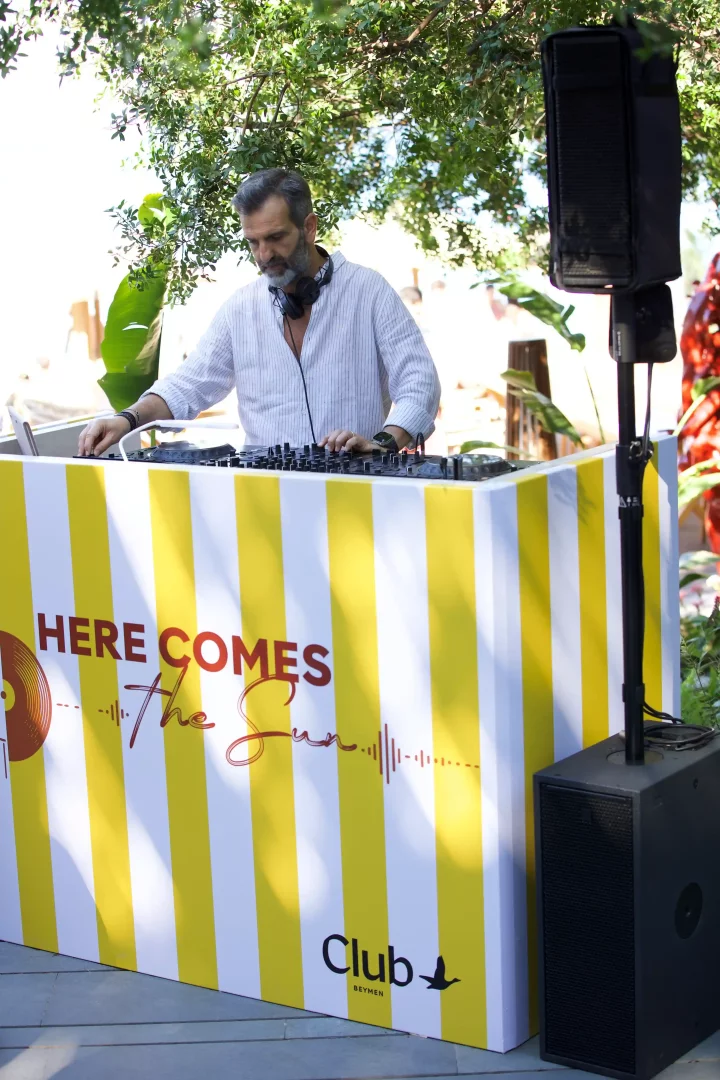 Bodrum'daki Beymen Club'ın altın yaz konseptli açık hava etkinliğinde, kulaklığı boynunda, sakallı bir DJ, üzerinde HERE COMES The Sun yazılı sarı beyaz çizgili kabinde müzik yaparken, yanında büyük bir hoparlör görülüyor.