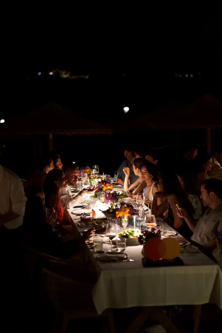 Gecenin karanlığında, şık bir Bodrum etkinliği için uzun bir masada toplanan konuklar, mum ışığı ve taze çiçeklerle süslenmiş ortamda yemek yiyip sohbet ederken, masanın ucunda görünen belirgin Mastercard logosu, etkinliğin Golden Summer temasına gönderme yapıyor gibi duruyor.