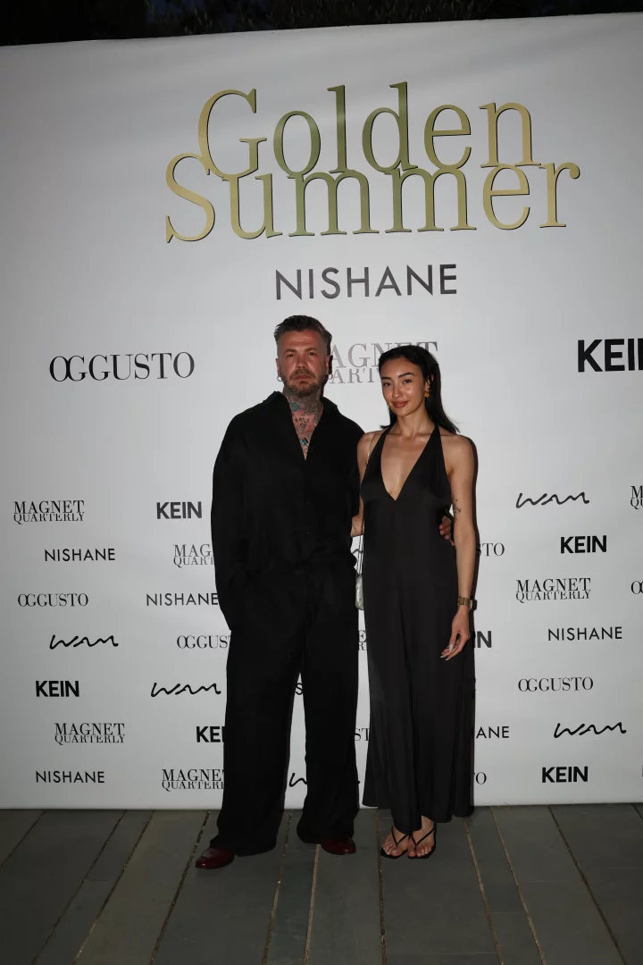 Golden Summer Bodrum etkinliği kapsamında, altın rengi Golden Summer yazısı ve Nishane, Oggusto gibi sponsor logolarıyla bezeli beyaz bir pano önünde, koyu renk, bol kesim kıyafetli dövmeli bir erkek ile askılı, derin V yakalı siyah tulum giymiş şık bir kadın birlikte poz veriyor.