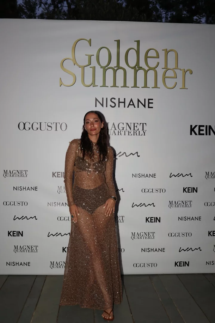 Etkinlikte objektife poz veren bir kadın, üzerinde parıltılı, şeffaf, bronz rengi uzun kollu bir elbise ve koyu renk bir mayo ile şık bir duruş sergiliyor; arka planda, bu özel Golden Summer etkinliğinin adını taşıyan büyük altın harfli yazı ve NISHANE, OGGUSTO gibi markaların logolarıyla süslenmiş beyaz bir pankart, Bodrum'daki bu yaz buluşmasının atmosferini belirginleştiriyor.