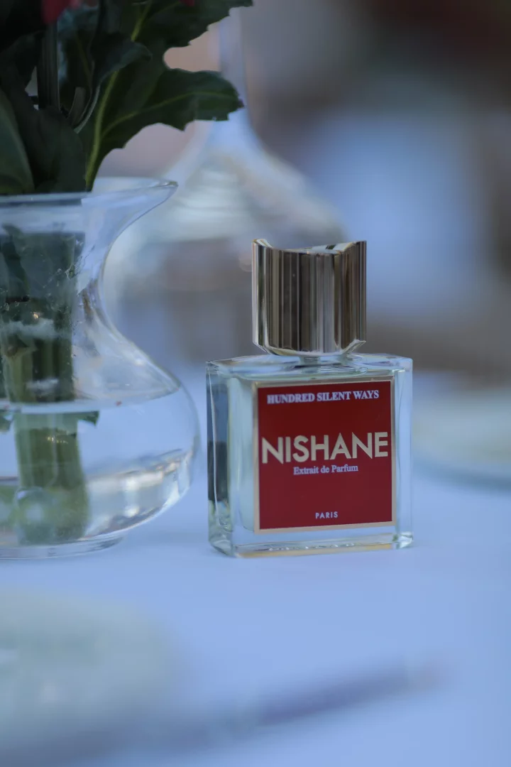 Aydınlık bir masa üzerinde, kırmızı etiketinde NISHANE ve HUNDRED SILENT WAYS Extrait de Parfum yazıları belirgin olan, şık metalik kapaklı cam bir parfüm şişesi, Altın Yaz Bodrum temasını çağrıştıran zarif bir kompozisyonun parçası olarak, solunda flu görünen yeşil yapraklı bir vazo ile konumlanmış.
