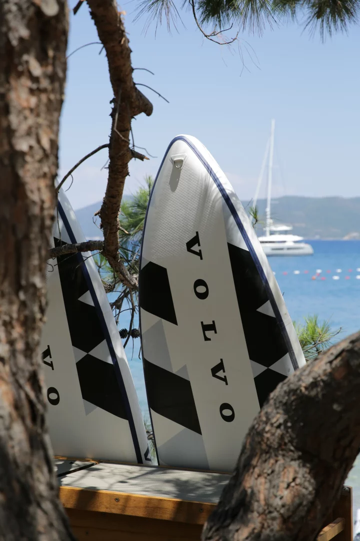 Çam ağaçlarının gövdeleri arasından beliren, üzerinde VOLVO yazılı iki beyaz paddleboard, Golden Summer Bodrum etkinliğinde denizin ritmini keşfetmek için beklerken, arka planda berrak Akdeniz suları ve açıkta demirlemiş bir yat görülüyor.