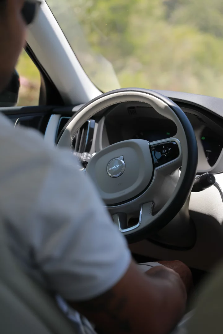 Sürücünün omzunun üzerinden çekilen bu görüntüde, güneşli bir Bodrum yaz gününde, direksiyon simidinde belirgin Volvo logosu ve kontrol düğmeleri bulunan modern bir aracın içinde, beyaz tişörtlü bir sürücünün hafifçe sağa dönük direksiyonu tuttuğu ve dışarıdaki bulanık yeşilliklerin ön camdan göründüğü an yakalanmış.