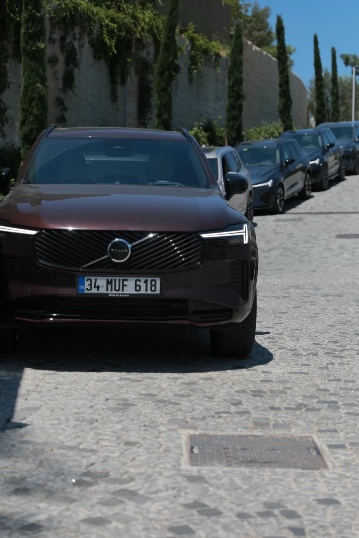 Önde park etmiş, 34 MUF 618 plakalı, koyu bordo rengi modern bir Volvo SUV, Bodrum'un güneşli bir yaz gününde taş döşeli yolda, arkasında düzenli bir sıra halinde bekleyen diğer aynı model Volvo araçlarla beraber, yeşilliklerle kaplı duvarın önünde şık bir görünüm sergiliyor.