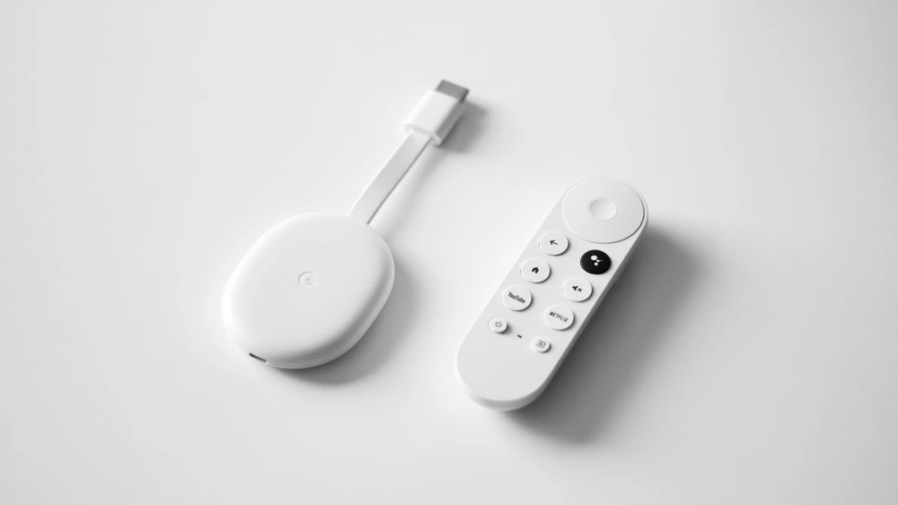 Beyaz bir Google Chromecast yayın cihazı ve ona ait kumanda, minimalist bir beyaz zeminde duruyor; oval tasarımlı Chromecast ile YouTube, Netflix ve Google Asistan tuşlarına sahip kumanda, sıradan televizyonları Smart TV'ye dönüştürme potansiyelini açıkça gösteriyor.