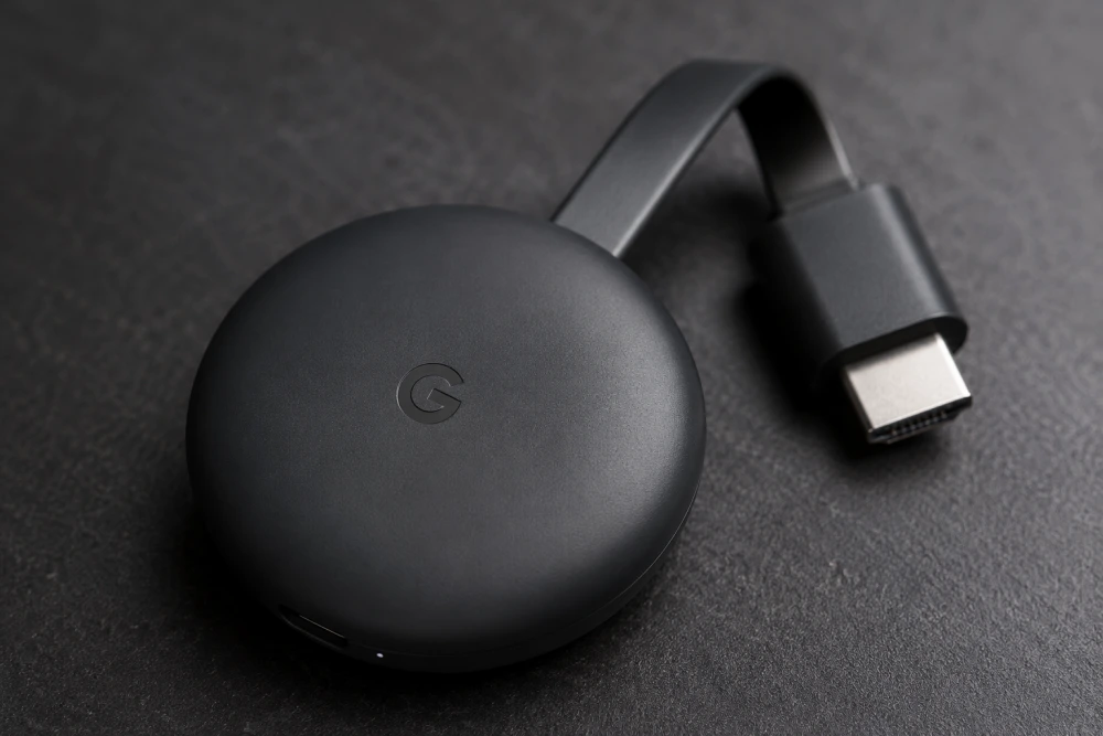 Koyu renkli, hafif dokulu bir yüzey üzerinde duran, üzerinde Google'ın sembolik G logosu bulunan siyah Google Chromecast cihazı, modern bir akıllı televizyon deneyimi sunmak için tasarlanmış HDMI bağlantı ucuyla dikkat çekiyor.