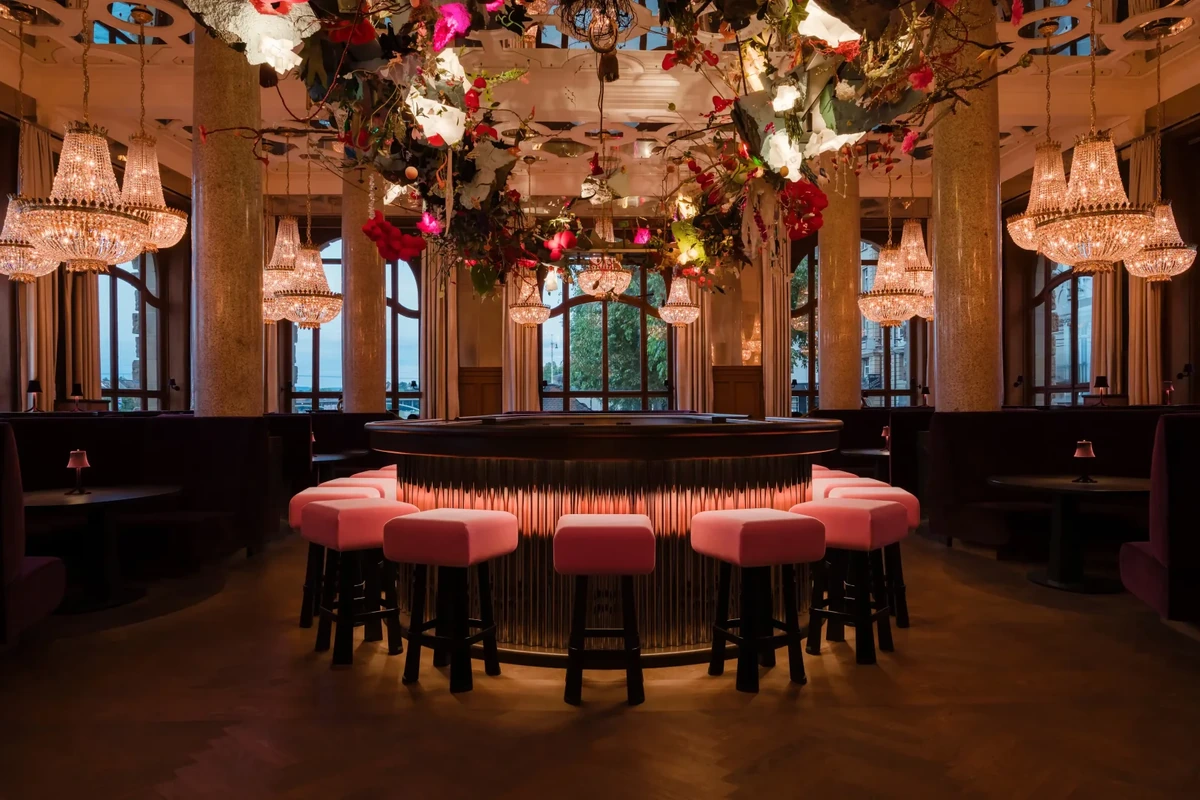 Grand Hotel Les Trois Rois bünyesindeki Banks Bar'da, altından sıcak turuncu ışık yayan oval bar tezgahını çevreleyen pembe minderli tabureler ve tavandan sarkan görkemli avizelerle ışıklandırılmış, canlı çiçek ve yeşilliklerle dolu devasa bir tavan dekorasyonuyla göz kamaştıran lüks ve şık bir iç mekan.