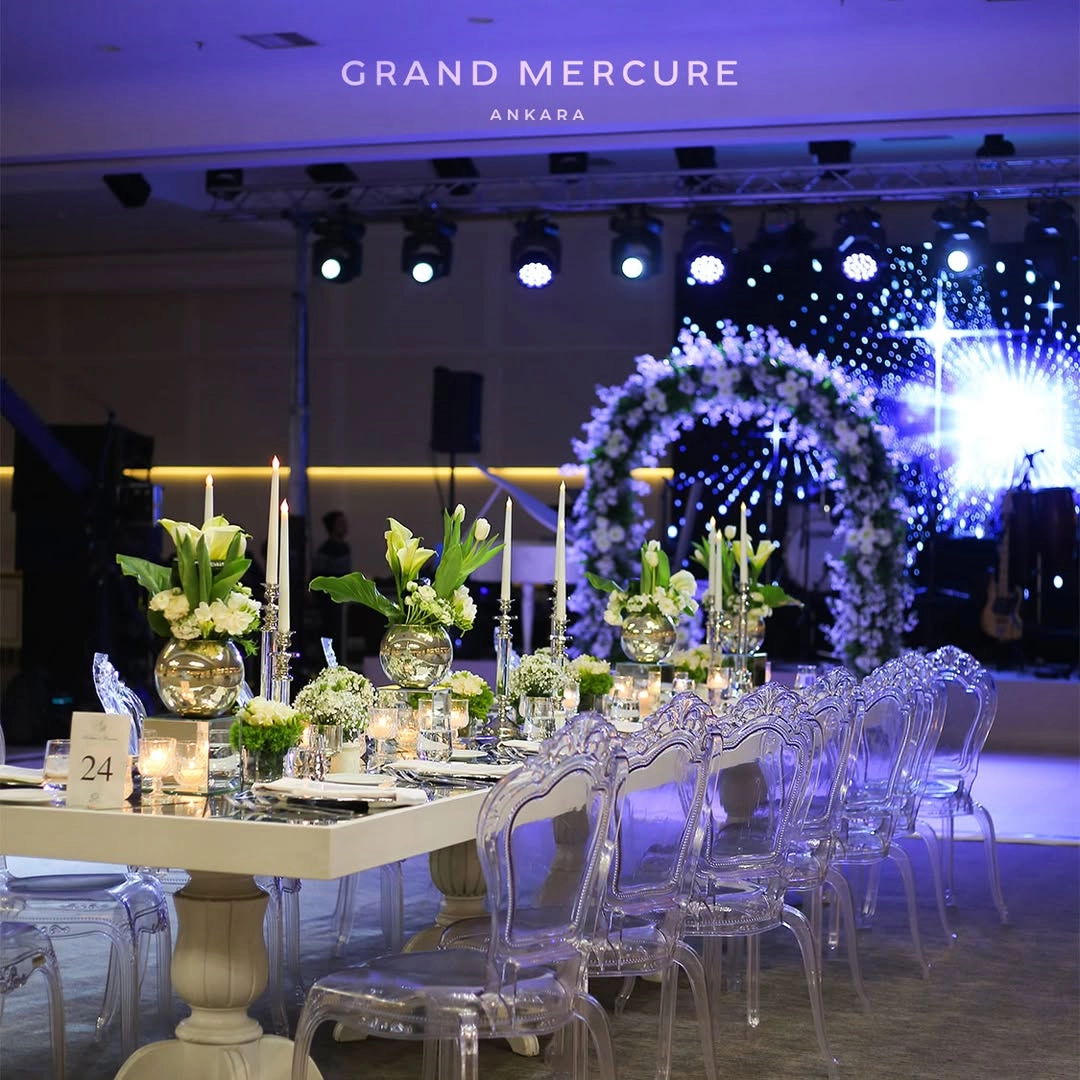 Ankara'daki gözde düğün mekânlarından Grand Mercure Ankara'da, şık beyaz ziyafet masası, modern şeffaf sandalyeler, beyaz çiçek aranjmanları, zarif mumlar ve arkada parıldayan led ekranın önünde duran görkemli çiçekli tak ile lüks ve modern bir düğün organizasyonu atmosferi yansıtılıyor.