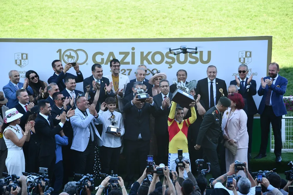 100. Gazi Koşusu ödül töreninde, sarı-kırmızı jokey forması giyen Halis Karataş büyük kupayı coşkuyla havaya kaldırırken, etrafındaki kalabalık alkışlarla kutluyor ve tören alanının üzerinde bir drone süzülüyor.