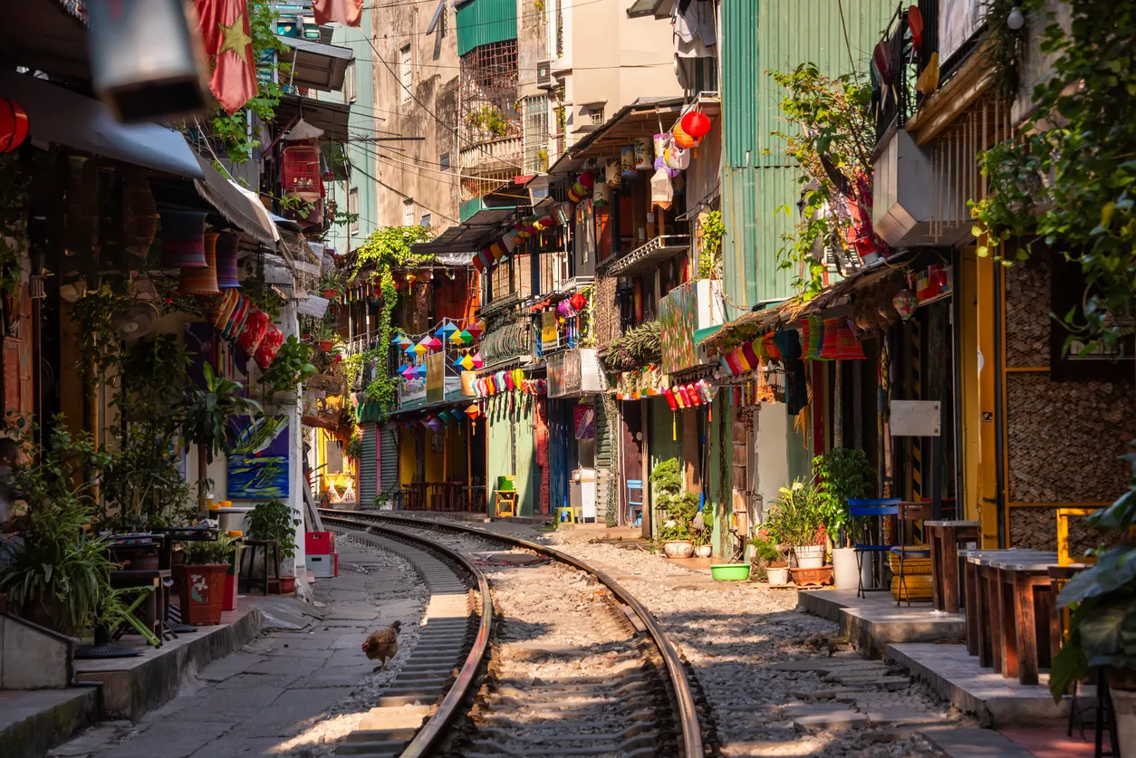 Hanoi'deki binaların rengarenk fenerler, çeşitli bitkiler ve tentelerle süslü olduğu, dükkanların sıralandığı dar bir sokakta, bir tavuğun kıvrılan demiryolu rayları üzerinde yürüdüğü, canlı ve otantik bir şehir manzarası.