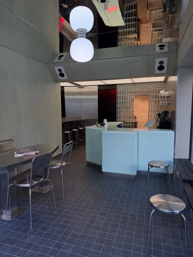 Harman Coffee Nişantaşı şubesinin minimalist ve modern iç mekanı, tavandan sarkan iki büyük küresel aydınlatma armatürü, açık mint yeşili tezgahı, metal masa ve tabureleri ile dikkat çekerken, koyu renkli karo zemini ve yansıtıcı tavanı şık bir atmosfer sunmaktadır.