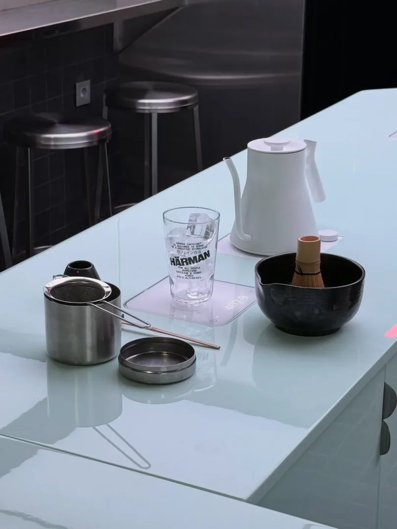 Modern ve minimalist bir cafe tezgahında, Harman Coffee markalı buzlu su bardağı, zarif beyaz bir kettle, matcha hazırlamak için kullanılan bambu çırpıcılı siyah kase ve paslanmaz çelik demleme ekipmanları düzenli bir şekilde sergileniyor.