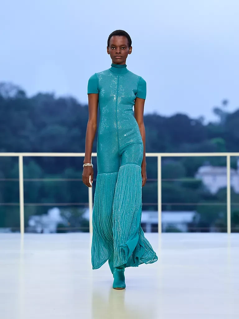 Hermès Resort 2027 defilesinde bir model, üst kısmı ışıltılı turkuaz payetlerle bezenmiş, bedeni saran ve alt kısmı geniş pilili, akışkan bir tulum ile uyumlu botlar giyerek podyumda zarifçe yürüyor; kıyafetin dans ve hareket özgürlüğünü vurgulayan silueti, açık mavi gökyüzü ve yeşil tepelerin oluşturduğu dingin Bel Air fonunda öne çıkıyor.