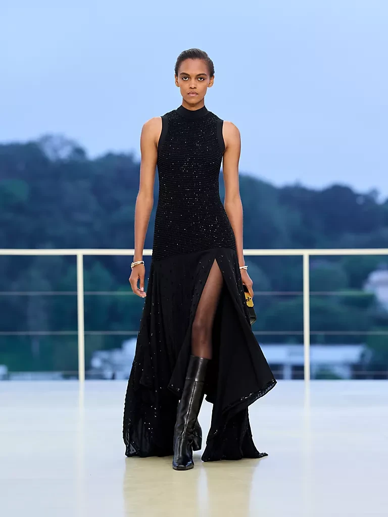 Hermès Resort 2027 defilesinden, parlak bir podyumda, yüksek yırtmaçlı, payet işlemeli siyah elbisesiyle dansın akışkanlığını yansıtan, zarif bir model, diz hizasındaki deri çizmeleri ve sade bileklikleriyle hareket ve özgürlük temasını vurguluyor.