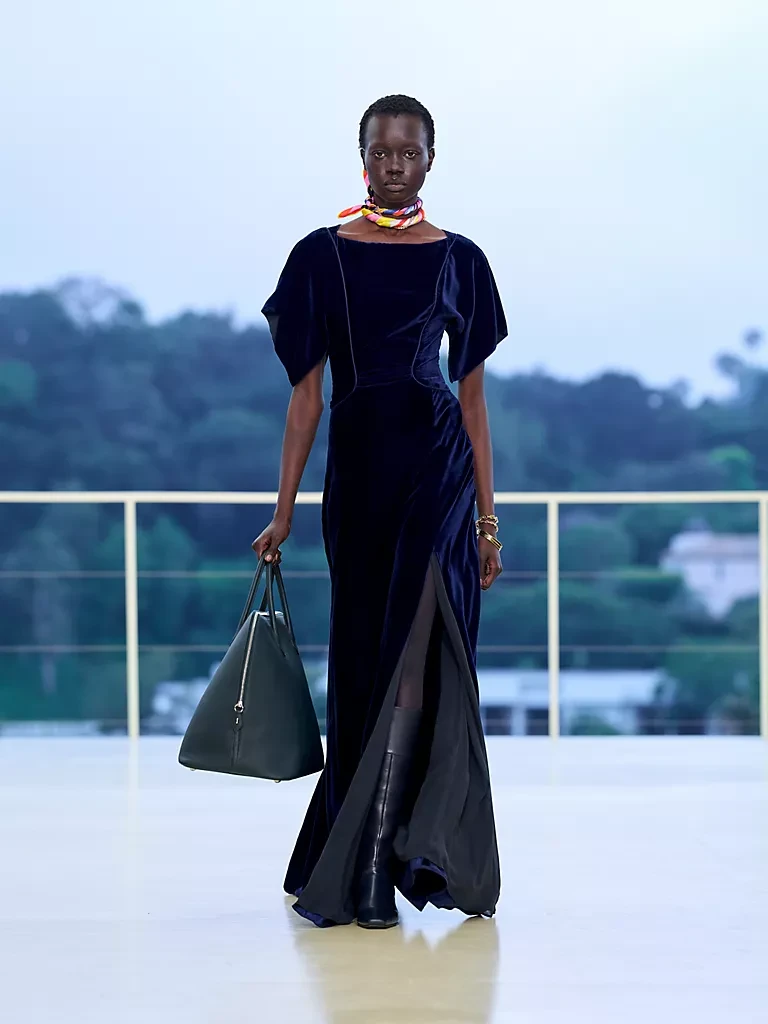 Hermès Resort 2027 defilesinden bir görünümde, koyu lacivert kadife, yırtmaçlı, akışkan bir elbiseyle yürüyen model; boynunda renkli bir fular, bileğinde altın bilezikler ve markanın yeni silüetlerinden biri olan, koyu yeşil, üçgen formunda büyük bir çantayı Bel Air'in gün batımı atmosferinde zarifçe taşıyor.
