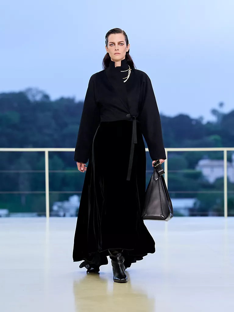 Hermès Resort 2027 koleksiyonundan, Bel Air tepelerinde gün batımına karşı podyumda yürüyen bir model, akışkan siyah kadife maksi etek, modern kesim sargı ceket ve ikonik tarzını yansıtan büyük siyah bir deri çanta ile güçlü ve zarif bir siluet sunuyor.