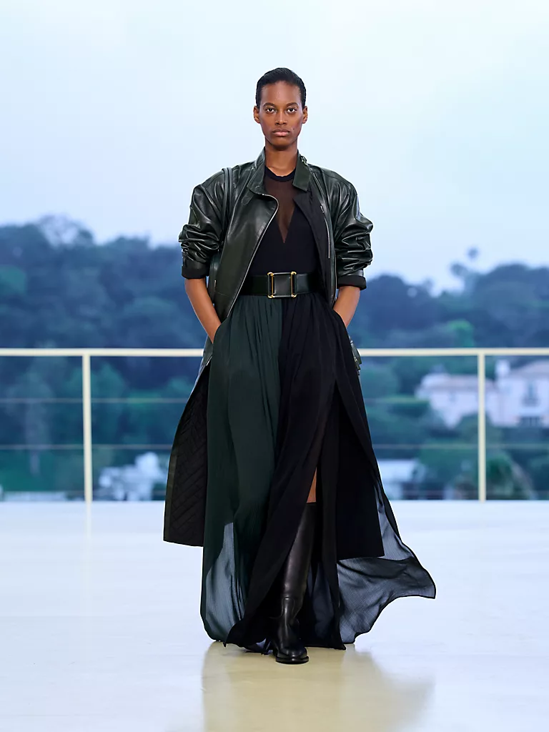 Los Angeles Bel Air tepelerindeki Hermès Resort 2027 defilesinde, koyu yeşil deri ceketi, transparan siyah üstü, geniş kemeri ve akışkan kesimli yeşil-siyah uzun eteğiyle podyumda özgürce yürüyen bir model, markanın deri işçiliğindeki yeni, daha yumuşak yaklaşımını sergiliyor.
