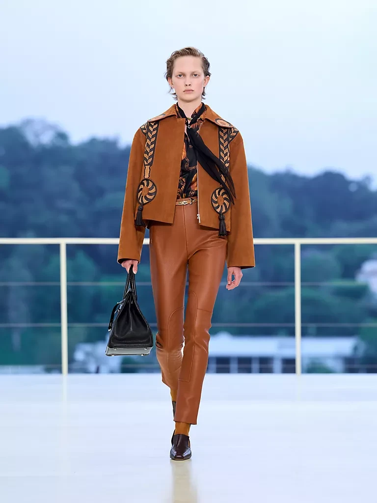 Hermès Resort 2027 Sonbahar-Kış koleksiyonundan bir model, Bel Air'in Los Angeles tepelerinde gün batımına karşı düzenlenen defilede, detaylı işlemelere sahip kahverengi süet ceketi, akışkan formdaki deri pantolonu, desenli bluzu ve zarif siyah çantasıyla yürüyerek markanın özgür ruhlu, güçlü deri işçiliğini vurguluyor.