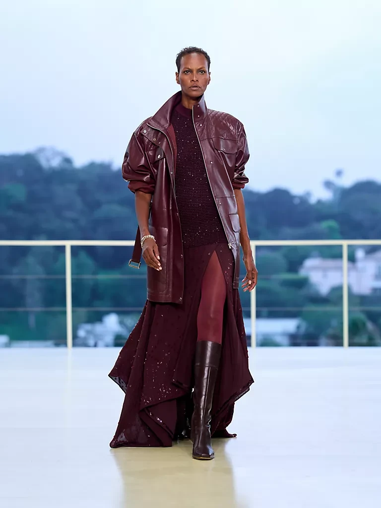 Yeni Hermès Resort 2027 koleksiyonundan, akışkan formuyla dikkat çeken, geniş yakalı ve omuzları kapitone bordo deri bir ceket, simli, derin yırtmaçlı dökümlü bordo bir elbise ve uyumlu dizüstü çizmeler giyen kısa saçlı, koyu tenli bir model, Los Angeles Bel Air tepelerindeki podyumda kendinden emin adımlarla ilerliyor.