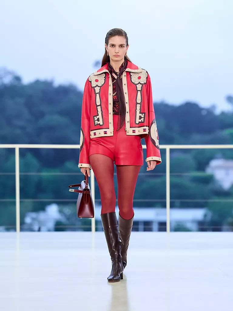 Hermès Resort 2027 Sonbahar-Kış koleksiyonundan, anahtar motifleriyle süslenmiş parlak kırmızı deri crop ceket ve şort takımı giyen bir model, bordo çanta ve diz üstü siyah çizmeleriyle gri gökyüzü altında podyumda yürüyor, markanın hareket ve özgürlükle buluşan akışkan deri işçiliğini vurguluyor.