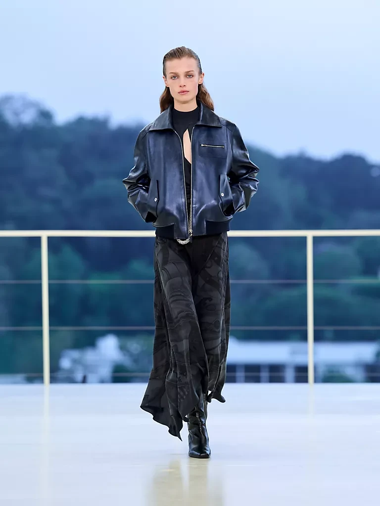 Model, Hermès Resort 2027 koleksiyonundan koyu renk parlak deri bomber ceket ve akışkan, desenli bir maksi etekle aydınlık bir podyumda yürürken, arkada bulanık ağaçlar ve açık renk bir gökyüzü görünmekte; bu görünüm markanın hareket, dans ve özgürlük duygusunu, akışkan deri ve zarafetle birleştirme vizyonunu vurguluyor.