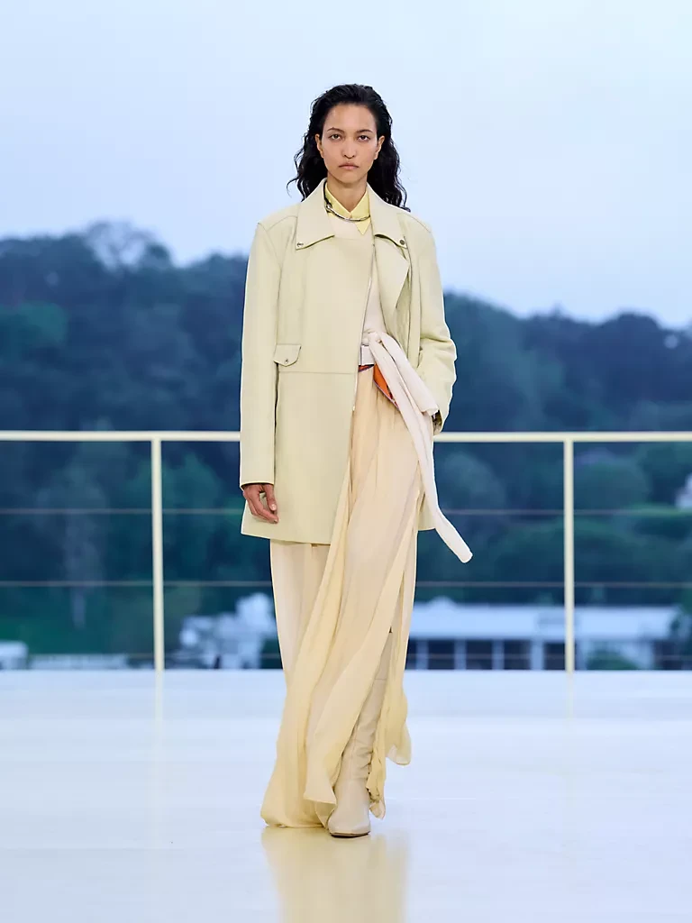 Hermès Resort 2027 defilesinden, tereyağı sarısı hafif bir ceket, açık sarı akışkan geniş paçalı pantolon ve beyaz çizmeler giyen bir model, Los Angeles Bel Air tepelerinde gün batımını yansıtan açık sarı pistte, hareket ve özgürlük temasını yansıtan bir görünümle kararlı adımlarla ilerliyor.