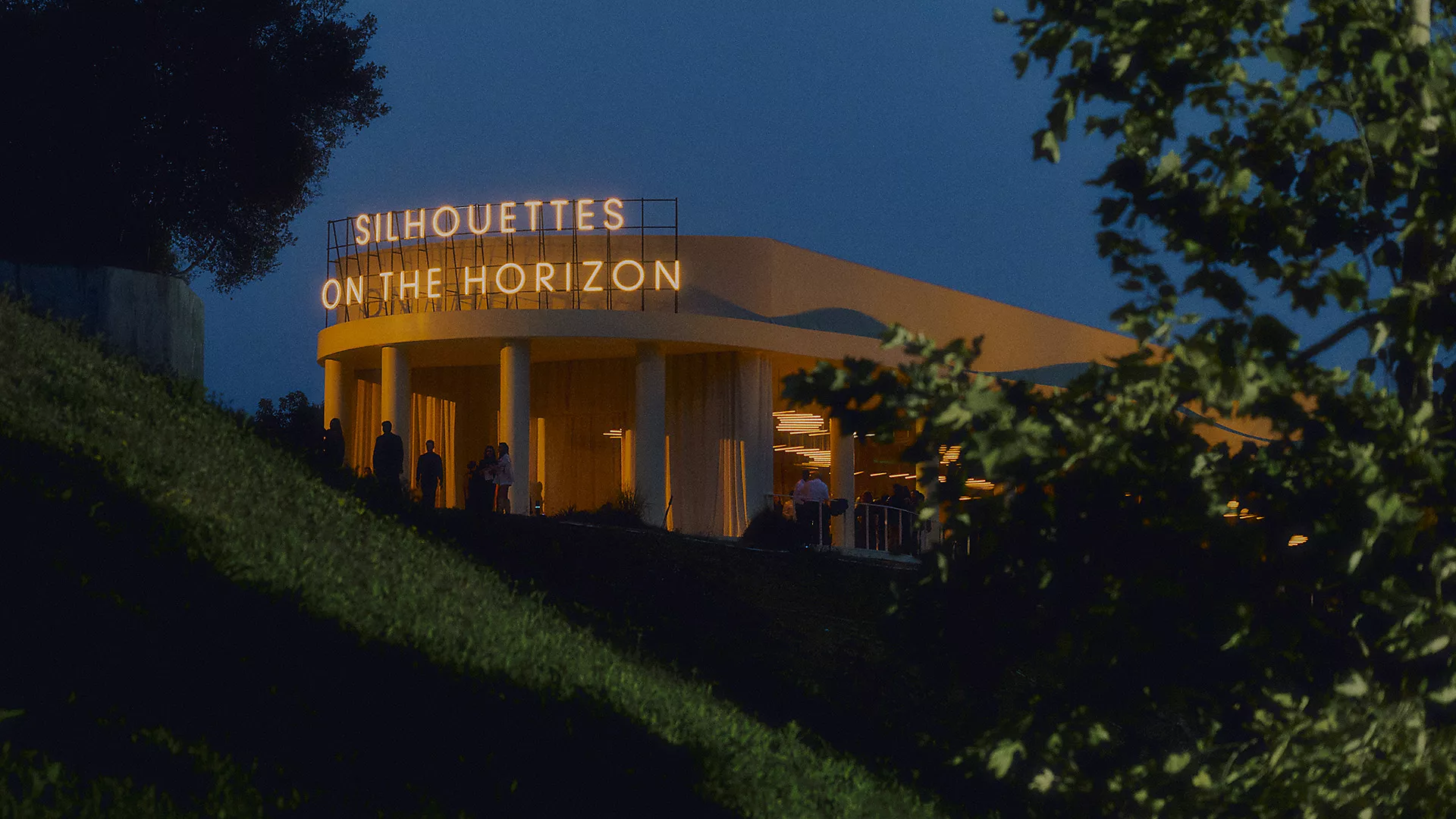 Hermès Resort 2027 Defilesinin Los Angeles'ın Bel Air tepelerinde, gecenin karanlığında ışık saçan, üzerinde Silhouettes on the Horizon yazısı parlayan modern pavyon mekânı ve etkinliğe katılan siluetler.