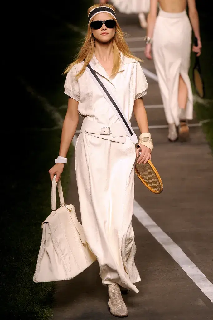 Sarışın, güneş gözlüklü ve saç bantlı bir model, Hermes İlkbahar 2010 hazır giyim koleksiyonundan, krem rengi, yakalı bir üst ve uzun etekten oluşan, bele oturan kemerli ve çapraz askılı bir kıyafetle, sol elinde büyük bir çanta ve sağ elinde ahşap bir tenis raketi taşıyarak, çimlerle çevrili, tenis kortu desenli podyumda kararlı adımlarla yürüyor.