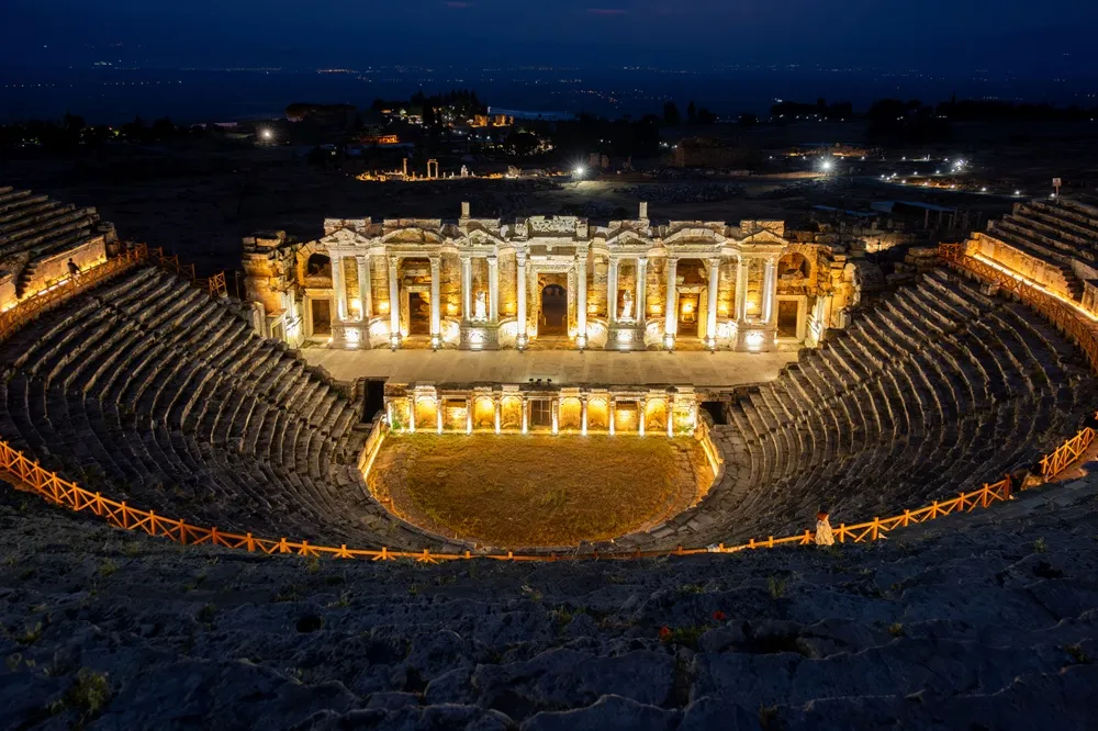 Hierapolis Antik Tiyatrosu'nun gece çekimi, Pamukkale'deki bu görkemli yapının ışıklandırılmış sahnesi ve basamaklı oturma alanlarıyla karanlık gökyüzü altında etkileyici bir şekilde yükseldiğini gösteriyor, uzakta şehrin ışıkları seçiliyor.