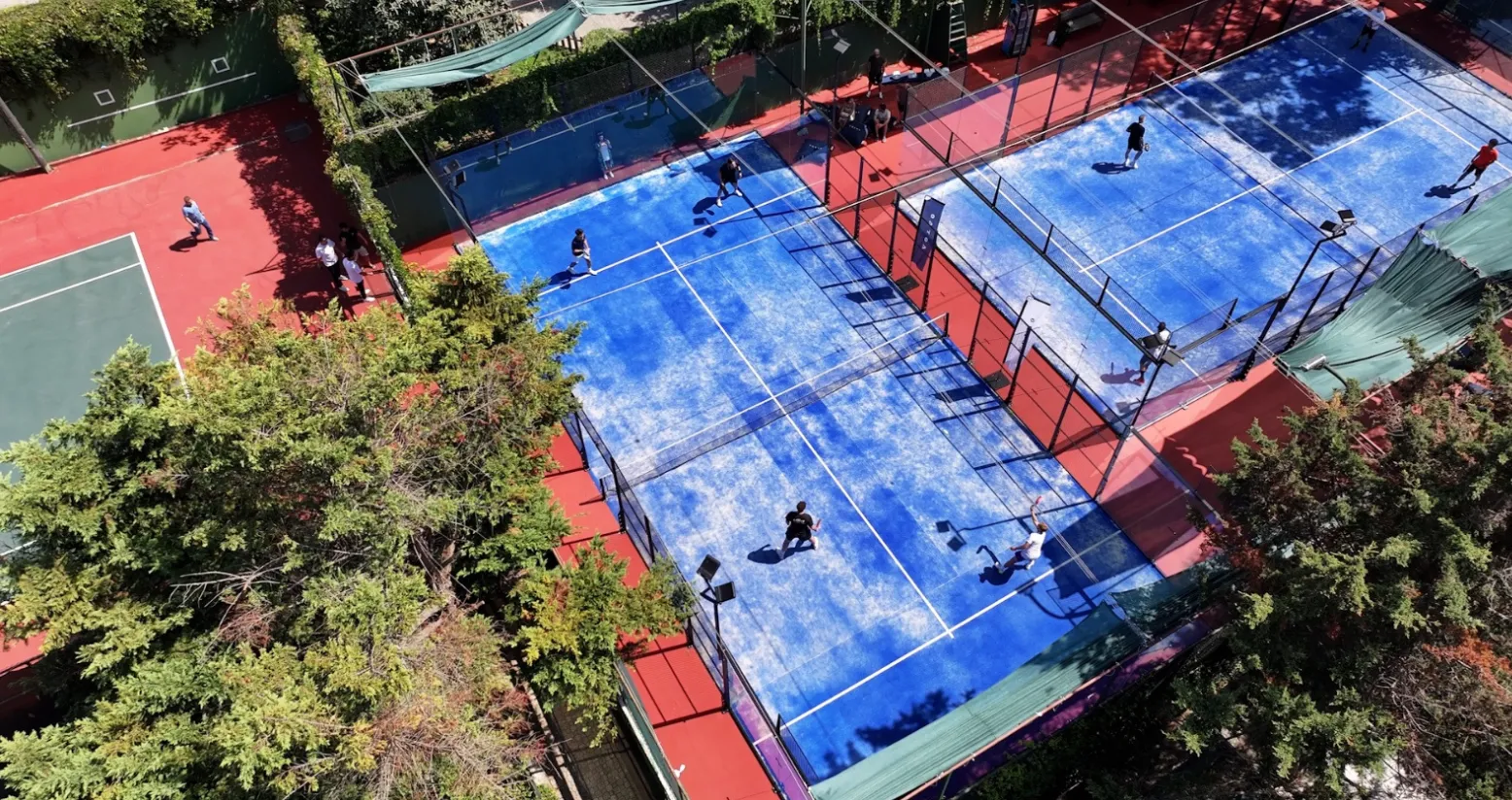 Hillside Etiler'deki açık hava spor tesisinden kuşbakışı çekilen bu fotoğraf, cam duvarlarla çevrili canlı mavi zeminli padel tenis kortlarında raketleriyle oynayan sporcuları ve kırmızı ile yeşil bir tenis kortunun yakınında bulunan diğer kişileri yemyeşil ağaçlar arasında gözler önüne seriyor.