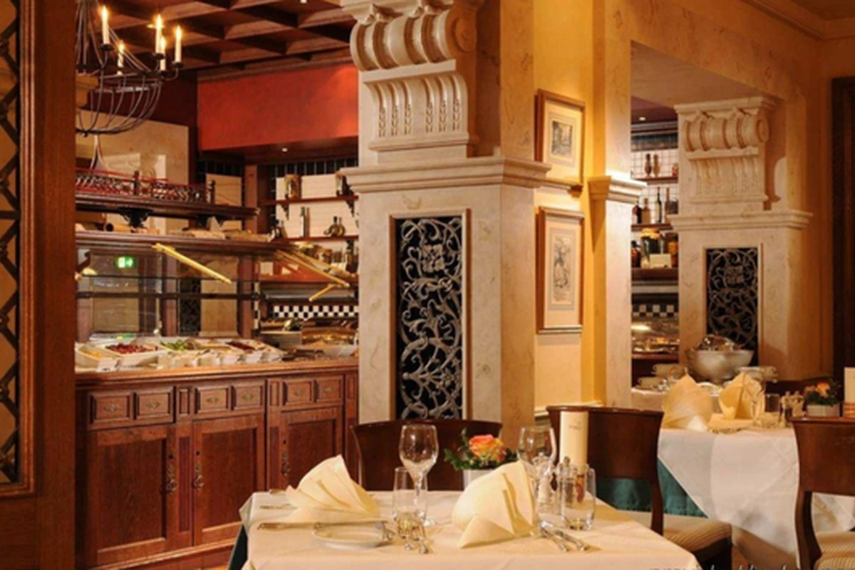 Hilton Basel restoranının sıcak ve davetkar atmosferini yansıtan bu görselde, özenle hazırlanmış masaların yanı sıra çeşitli lezzetlerin sunulduğu ahşap büfe alanı ve mekanın şıklığını artıran oyma detaylı dekoratif sütunlar dikkat çekiyor.