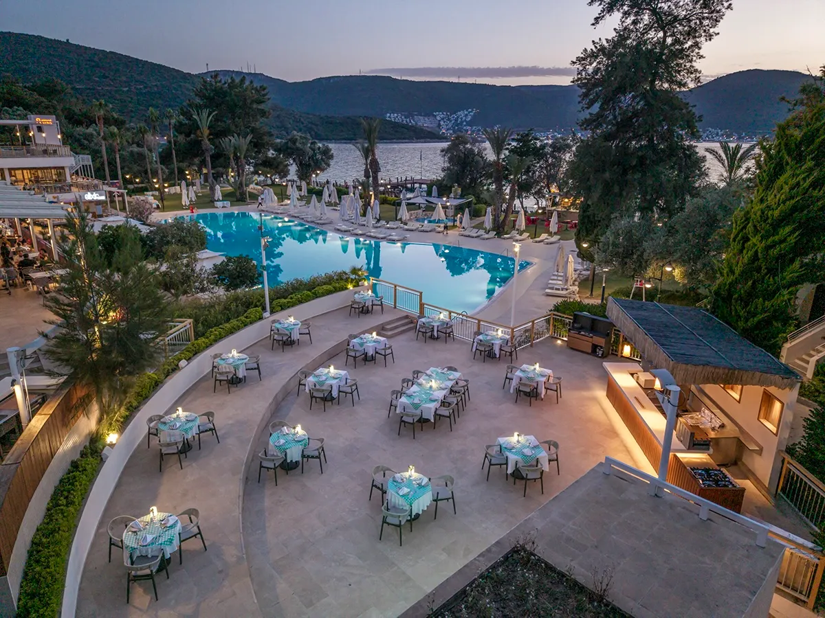 Gün batımında Bodrum'daki Hilton Bodrum Işıl Club Ultra All Inclusive Resort'un ışıltılı büyük yüzme havuzu ve Akdeniz manzaralı açık hava restoranının yer aldığı, evcil hayvan dostu bir tatil için davetkar bir atmosfer sunan geniş bir teras.