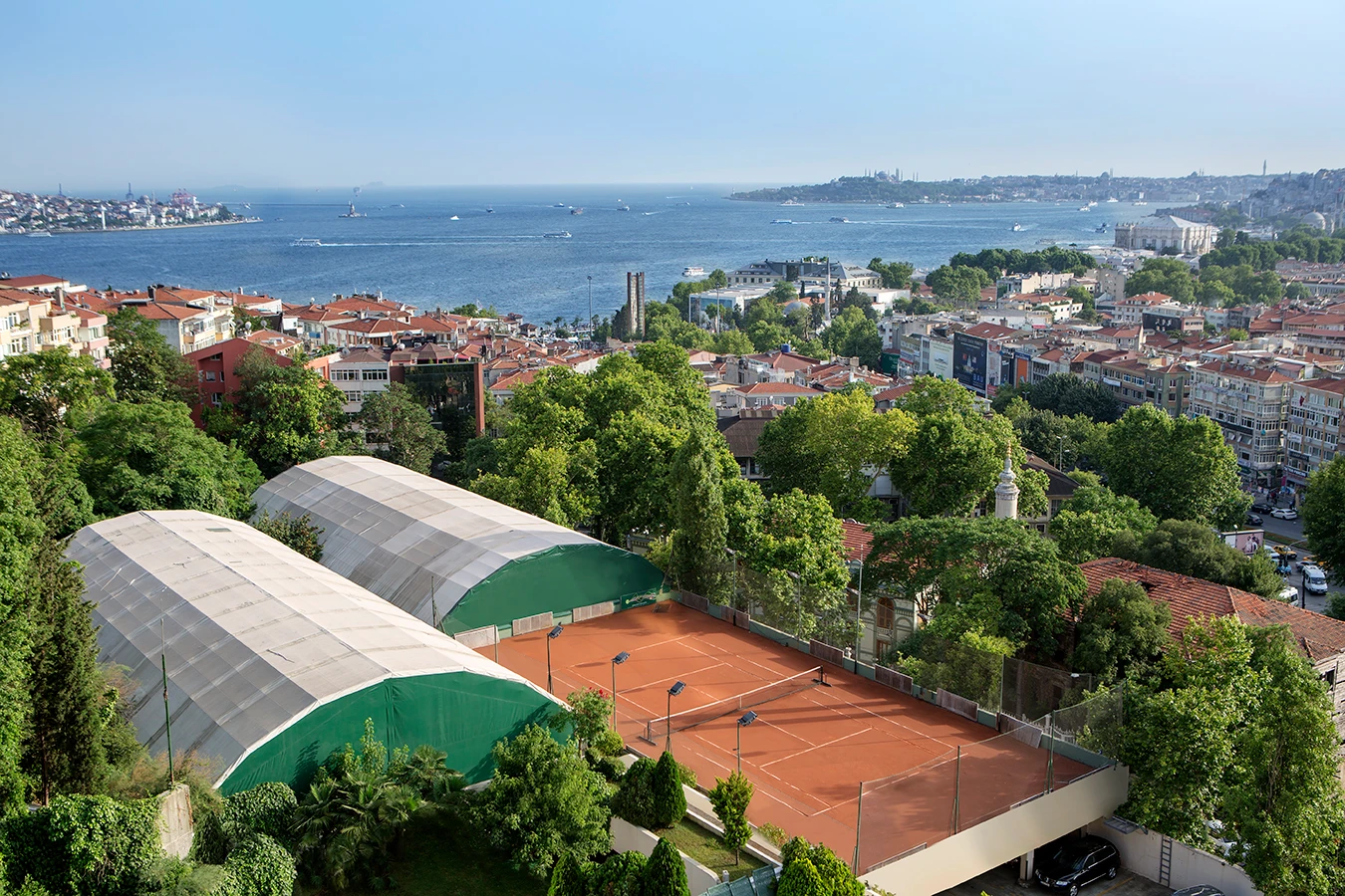 İstanbul Boğazı'nın eşsiz maviliği ve yemyeşil ağaçlarla çevrili bir alanda yer alan Hilton Conrad Tenis Kulübü'ne ait açık ve iki kapalı kort, şehir silüetiyle birlikte güneşli bir günde yukarıdan görülüyor.