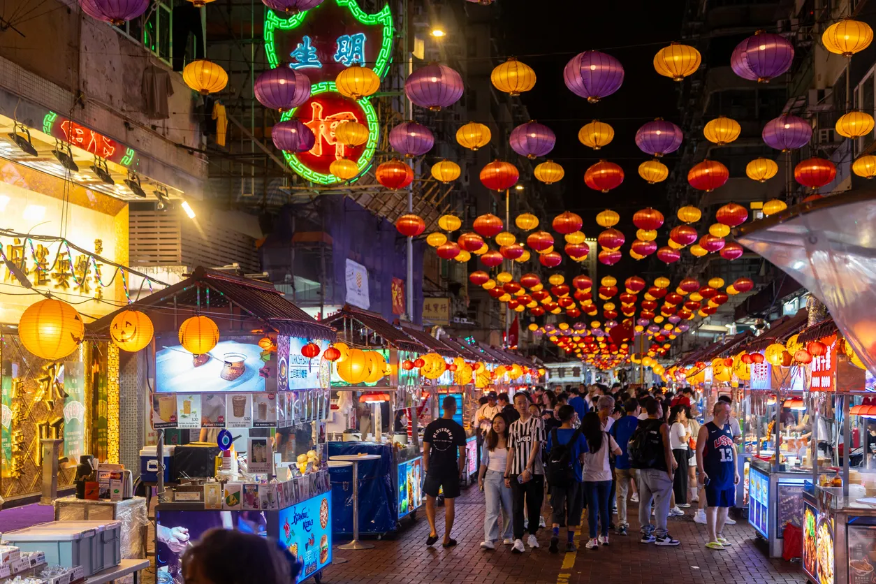 Hong Kong'da, gece vakti sokakları aydınlatan sarı, mor, kırmızı ve turuncu rengarenk fenerlerle süslenmiş, çeşitli yiyecek ve ürünler sunan tezgahlar arasında dolaşan kalabalıklarla dolu canlı bir gece pazarı manzarası.