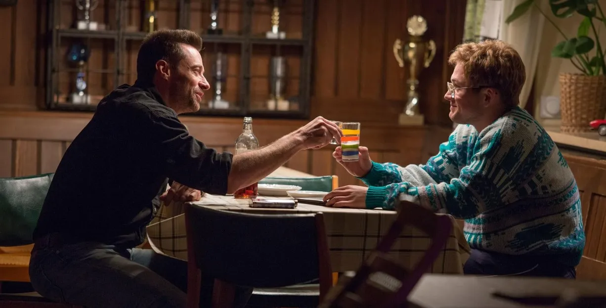 Hugh Jackman'ın yer aldığı 'Eddie the Eagle' filminden bir sahnede, Jackman'ın canlandırdığı içkiye düşkün eski şampiyon Bronson Peary, gözlüklü, renkli bir kazak giymiş, neşeli görünümlü sporcu Eddie Edwards rolündeki Taron Egerton ile ahşap bir barda kadeh tokuşturarak samimi bir an paylaşıyor; arka planda görünen kupalar ve spor temalı detaylar, bu Hugh Jackman filmleri arasında yer alan ilham verici spor dramasının atmosferini güçlendiriyor.