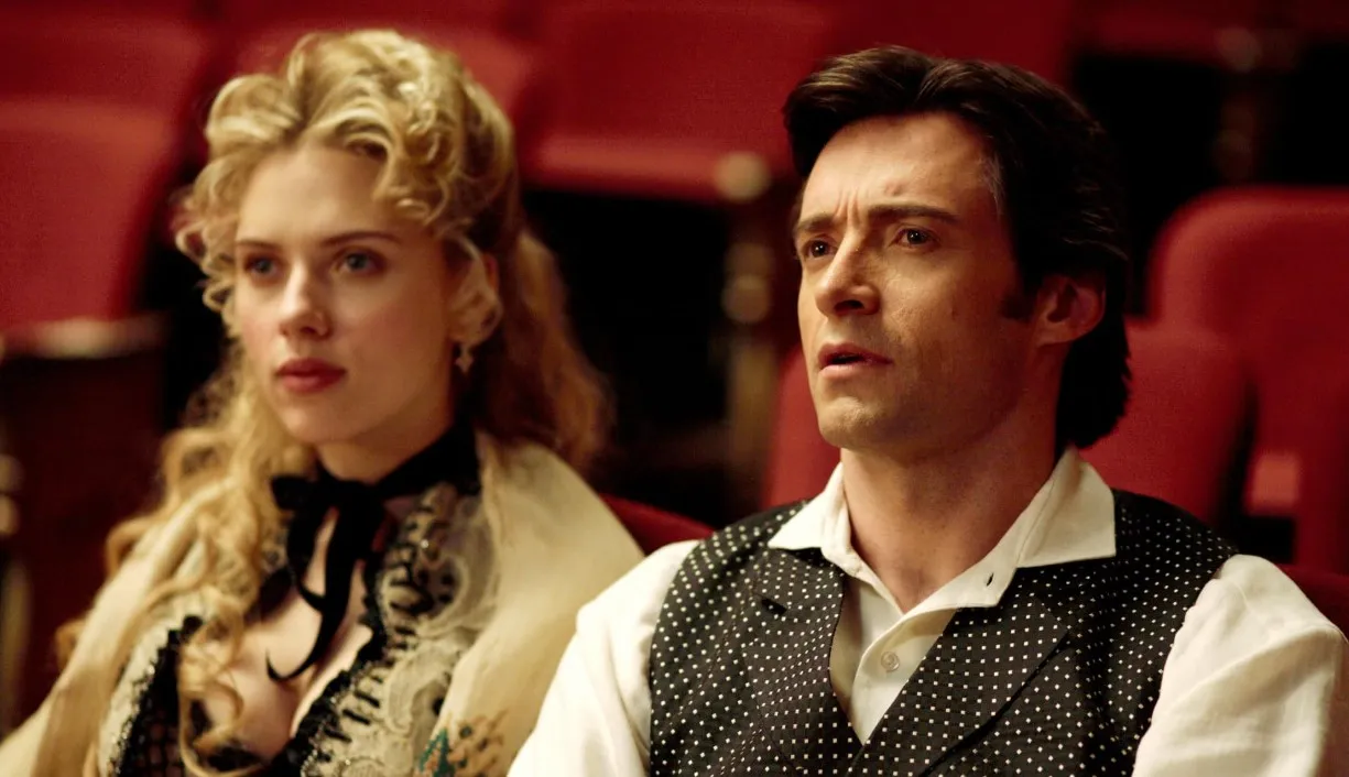 Hugh Jackman filmleri listesindeki Christopher Nolan imzalı The Prestige filminden bir karede, Viktorya dönemi kıyafetleri içindeki Hugh Jackman ve Scarlett Johansson, kırmızı tiyatro koltuklarında oturmuş, meraklı ve gergin bir ifadeyle sahneyi izliyorlar.