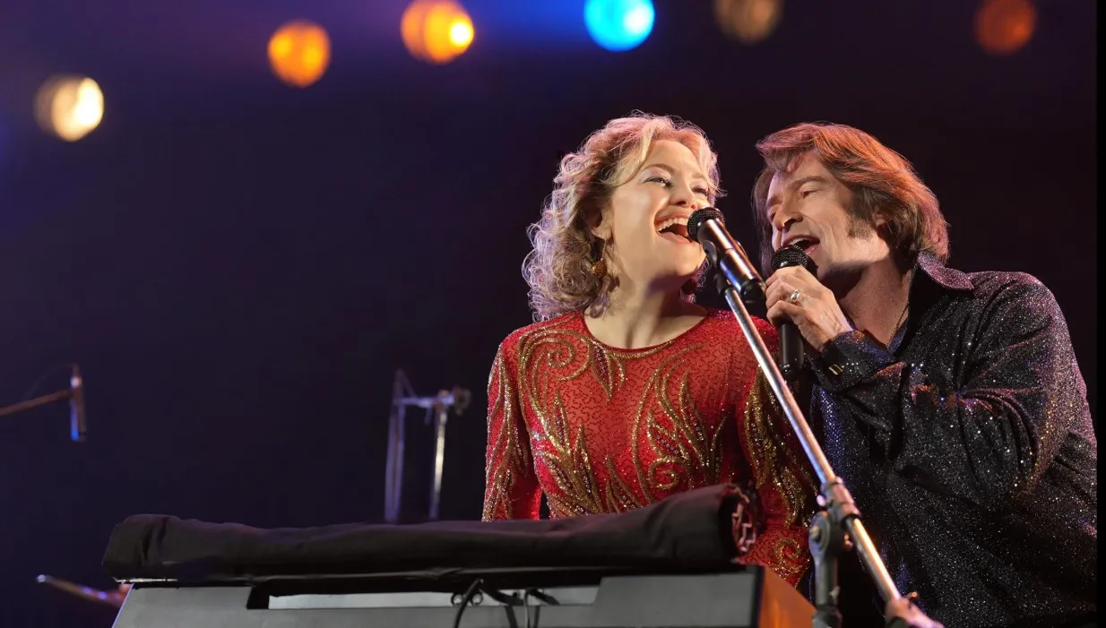 Song Sung Blue filminden bir karede, Neil Diamond tribute grubu üyeleri Mike ve Claire Sardina'yı canlandıran Hugh Jackman ve Kate Hudson, pullu sahne kostümleriyle coşkulu bir şekilde mikrofonlara şarkı söylüyor, karanlık sahne arka planında renkli ışıklar parlıyor.