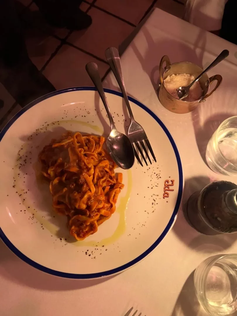 Ida London restoranında beyaz masa örtüsü üzerinde servis edilen, kırmızı soslu ve zeytinyağlı tagliatelle makarnanın olduğu tabakta bir kaşık ve çatal duruyor; tabağın kenarında kırmızı Ida yazısı, yanında bakır bir kapta rendelenmiş parmesan peyniri ve su bardakları dikkat çekiyor.