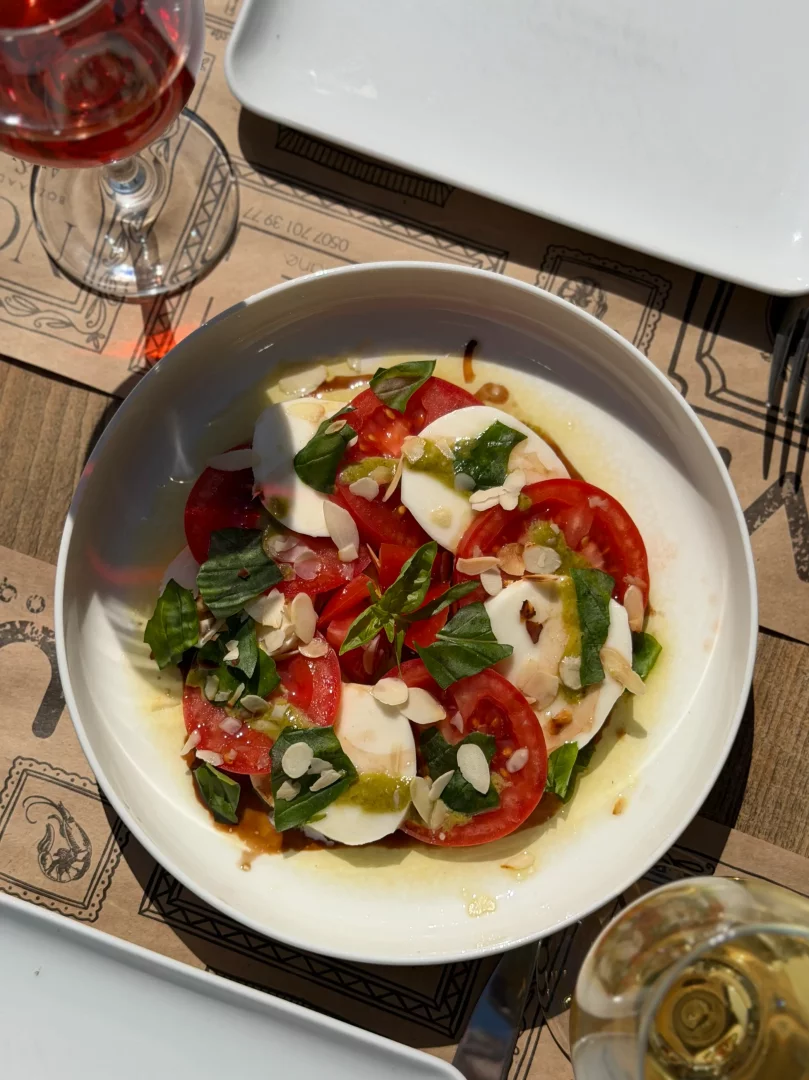 Güneşli bir masada, kahverengi desenli bir servis altlığının üzerinde parlak beyaz bir tabakta sunulan, dilimlenmiş taze domates, mozzarella, fesleğen yaprakları, badem pulları ile zeytinyağı, balzamik ve pesto soslarıyla zenginleştirilmiş Caprese salatası, iki kadeh şarap eşliğinde keyifli bir öğünü sergiliyor.