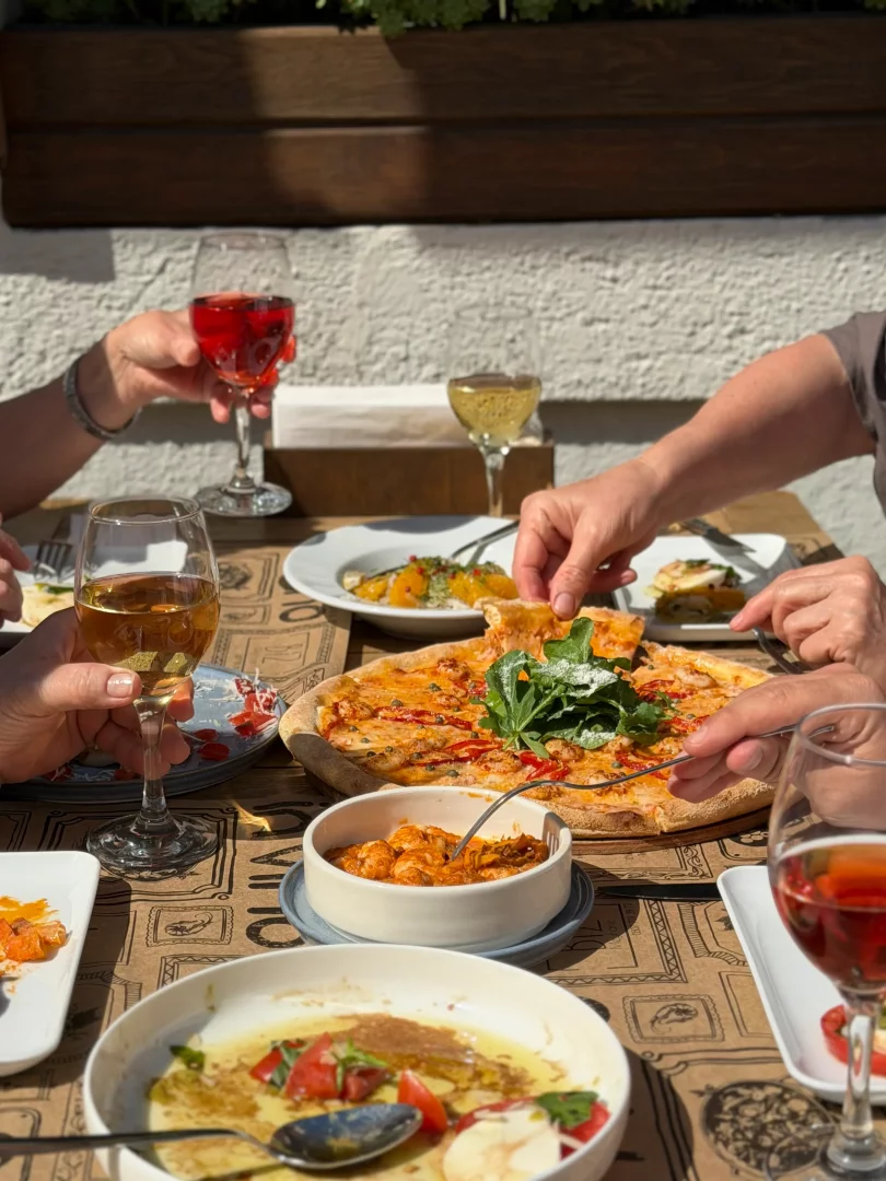 Güneşli bir ortamda, dört kişinin şarap kadehleri eşliğinde, roka ve karidesli görünen büyük bir pizzanın ve zeytinyağlı domates, burrata gibi çeşitli mezelerin bulunduğu bir masada keyifli bir akşam yemeği paylaştığı anı gösteren yakın çekim bir kare.