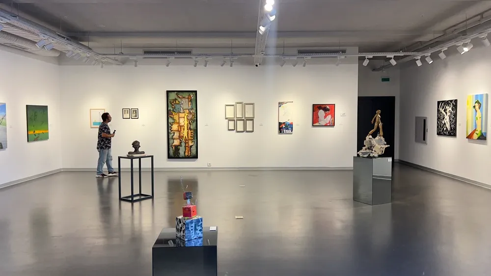Modern bir sanat galerisinin geniş ve aydınlık salonunda, beyaz duvarlarda farklı tarzlarda tablolar ve parlak gri zemindeki kaideler üzerinde heykeller yer alıyor; bir kişi sol taraftaki yeşil tonlardaki bir eseri dikkatle incelerken, bu sahne Bodrum yaz sergileri kapsamında sunulan zengin sanatsal seçkiyi gözler önüne seriyor.