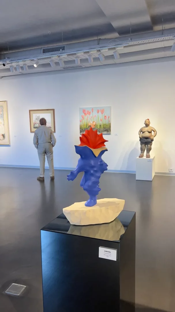 Inspera Bodrum'da düzenlenen bir sanat sergisinde, ön planda koyu mavi ve kırmızı tonlarda soyut bir heykel siyah bir kaide üzerinde sergilenirken, arkada bir ziyaretçi duvardaki tabloya bakıyor ve galeride başka heykeller ile tablolar da görülüyor.