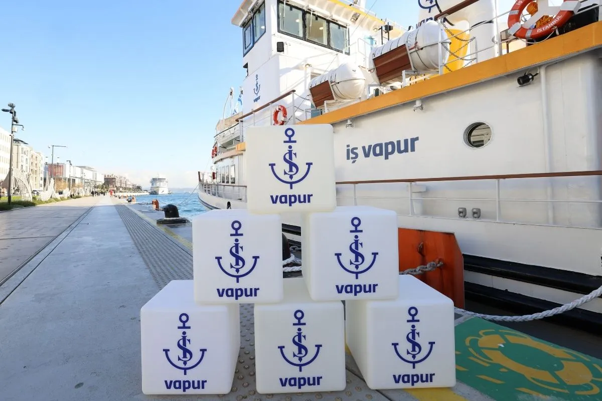 Galataport iskelesinde demirlemiş beyaz İş Vapur'un önünde, mavi çapa ve vapur logosu taşıyan altı beyaz küpün sergilendiği bu canlı görüntü, İş Sanat'ın yetişkinler ve çocuklara yönelik İş Vapur etkinlikleri için sanatsal programların başladığını müjdeliyor.