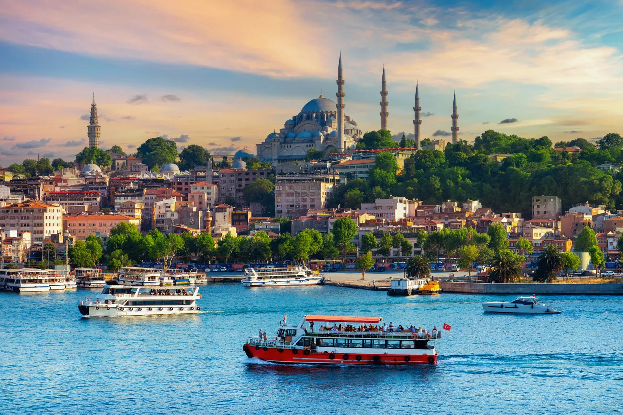 Gün batımının kızıl ve turuncu tonlarında parlayan gökyüzünün altında, İstanbul'un tarihi yarımadası üzerinde yükselen heybetli Süleymaniye Camii'nin minareleri ve kubbeli yapısı belirginleşirken, Haliç'in masmavi sularında yüzen turist tekneleri ve vapurlar bu büyüleyici şehir manzarasını tamamlıyor.