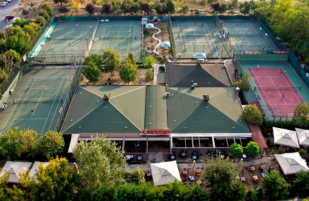 Ağaçlarla çevrili, yeşil ve kırmızı zeminli birçok tenis kortunun aktif olarak kullanıldığı, geniş bir kulüp binası ve açık hava oturma alanlarının yer aldığı İstanbul Tenis Spor Kulübü'nün kuşbakışı görüntüsü.
