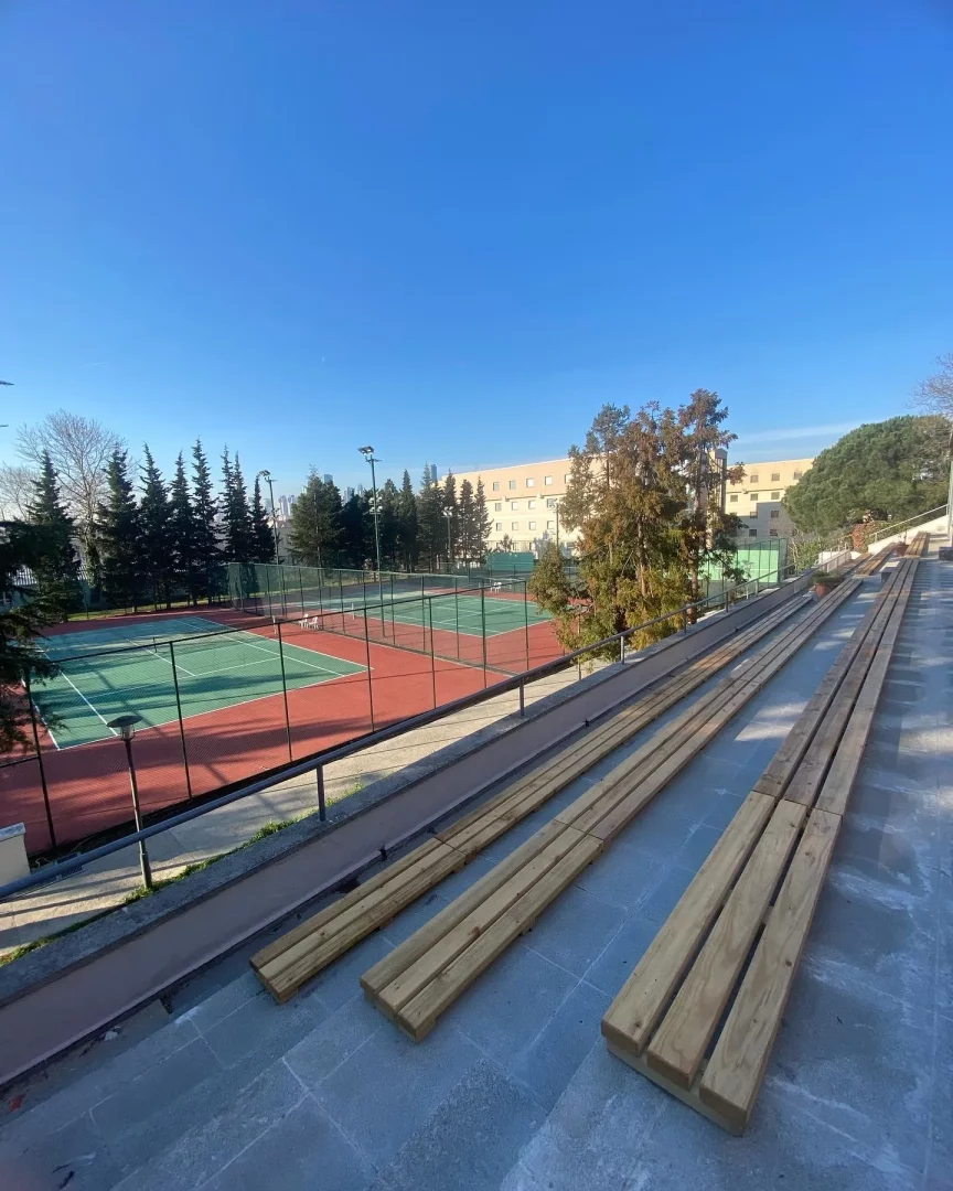 ITU Ayazağa Kampüsü'nde, parlak mavi bir gökyüzünün altında uzanan, yeşil ve kırmızı yüzeyli iki tenis kortu, çevresi tel örgülerle çevrili, soluk sarı bir binanın ve çeşitli ağaçların kuşattığı bir manzarada, ön planda ise seyirci alanı için hazırlanmış uzun ahşap oturma elemanları belirgin bir şekilde görülmektedir.