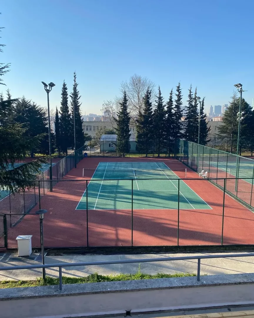 İTÜ Ayazağa kampüsünde yer alan, etrafı yüksek ağaçlar ve tel örgülerle çevrili, kırmızı zeminli ve yeşil oyun alanına sahip iki açık hava tenis kortu, berrak mavi gökyüzünün altında ve uzaktaki şehir manzarasının eşliğinde gün ışığında huzurlu bir görünüm sunuyor.